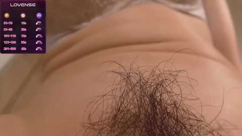 Video of a close-up view of a woman's lower abdomen, showing a dense, dark pubic hair patch and a glimpse of her white bra strap. Background includes a chart displaying menstrual cycle data.