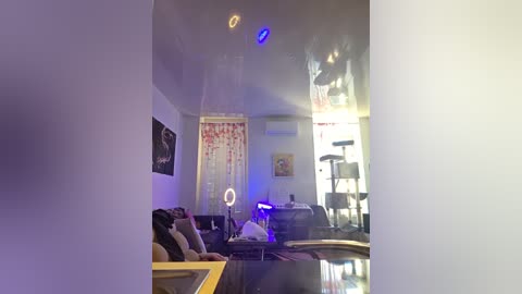 Video of a modern, dimly lit living room with white walls, a black sofa, a blue neon light, a cat tree, and a large, colorful tapestry hanging from the ceiling.