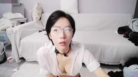 Media: Video of a young East Asian woman with short black hair, wearing glasses and a white blouse, kneeling in a messy room with white furniture.