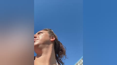 Video of a Caucasian woman with long brown hair, gazing upwards against a vivid blue sky. Her skin is tanned and she wears a white top.