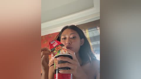 Media: A video of an Asian woman with long dark hair, wearing only a red bra, eating instant noodles from a bowl. Background features a beige wall and a window with yellow curtains.