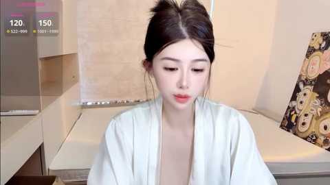 Video of a young Asian woman with fair skin, wearing a light robe, sitting in a modern bathroom with beige tiles and a colorful cartoon poster in the background.