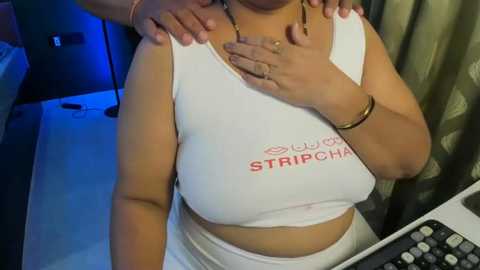 Video of a woman in a white sports bra with \"STRIP CHAT\" text, showing her medium-sized breasts, medium skin tone, and wearing a gold bracelet. Background includes a keyboard and a dark curtain.