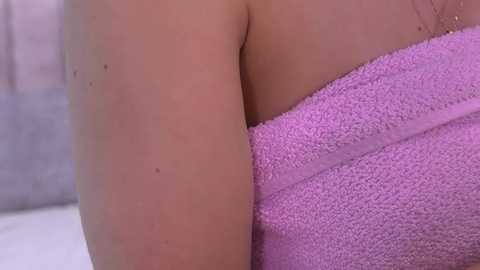 A close-up video of a light-skinned person's upper arm and shoulder wrapped in a fluffy, pink towel, with a blurred, light-colored background.