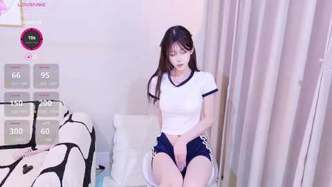 A video of an East Asian woman with long dark hair, wearing a white crop top and black shorts, sitting on a bed in a white room.