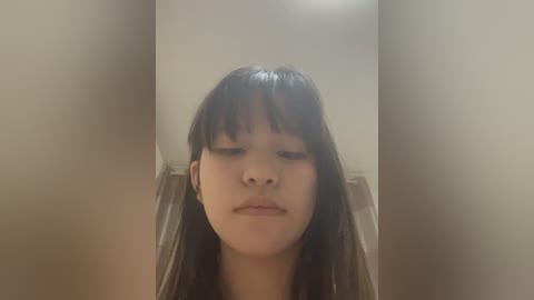 Video of a young Asian woman with long, straight black hair and light skin, wearing a neutral expression. She stands in a dimly lit, beige-colored room, likely a bathroom, with blurred background.