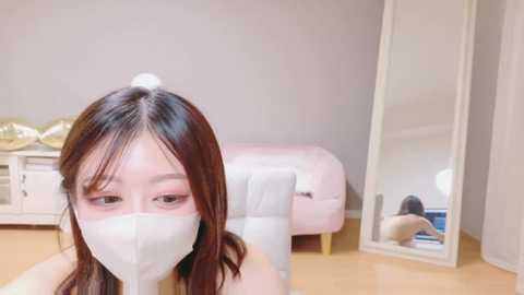 A video of an Asian woman with long, dark hair wearing a white mask, sitting on a pink sofa in a minimalist, light-colored room with a mirror reflecting her and a white wardrobe.
