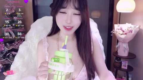 A young Asian woman with long black hair, wearing a white fur coat and pink lingerie, holds a green drink with a straw. Background includes a vase of flowers and a lamp.