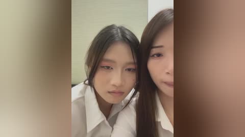 Video of two Asian women with fair skin, long black hair, and makeup. They wear white shirts, one resting her head on the other's shoulder. Background shows a beige, ribbed wall.