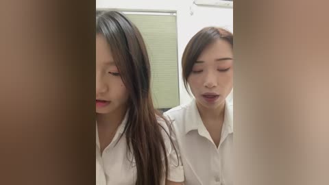 Video of two Asian women with long dark hair in white shirts, eyes closed, possibly asleep, in a dimly lit room with a closed window.