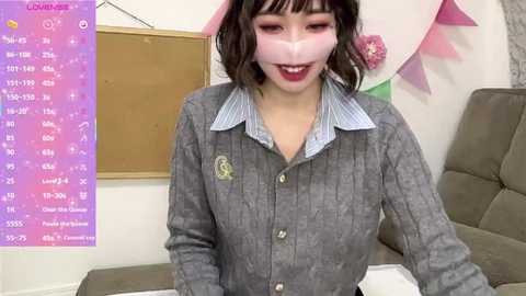 Video of an East Asian woman with shoulder-length dark hair and fair skin, wearing a grey cardigan over a light blue collared shirt, smiling. Background includes a pink and green wall with a calendar.