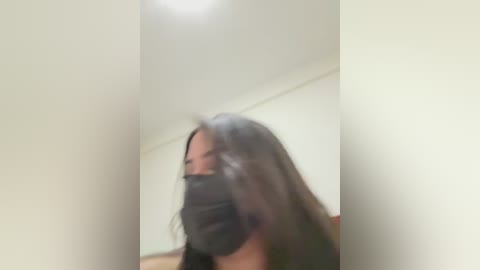 A blurry video of a person with long, dark hair and a black mask, standing in a narrow, white-walled corridor with a ceiling light.