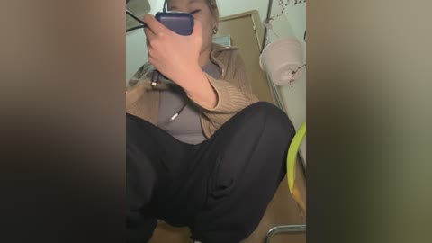 Video of a person sitting, holding a smartphone, wearing a beige jacket, black pants, and a gray shirt, in a dimly lit room with a white lamp, wall, and door.