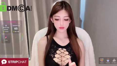 A video of an Asian woman with long brown hair, fair skin, and a black lace bodysuit with cutout details. She is seated in a white chair, in a room with beige curtains.