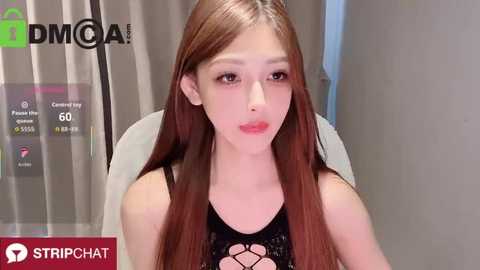 A video of a young Asian woman with long, straight brown hair, wearing a black lace top, sitting in front of a computer screen displaying a DMCa logo and streaming chat.