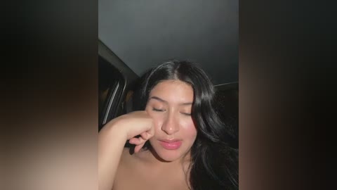 Video of a young woman with long, dark hair, light brown skin, and closed eyes, resting her chin on her hand. The background is dimly lit with dark tones, suggesting an indoor setting.