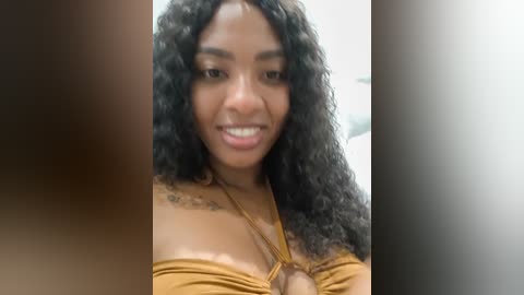 Video of a smiling young Black woman with curly hair, wearing a mustard-yellow off-shoulder top, with a blurred background.