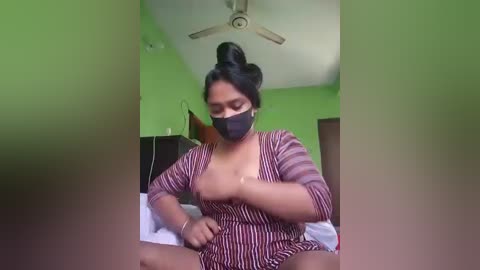 Media: Video of a South Asian woman with medium brown skin, wearing a black face mask, striped dress, and hair in a bun, adjusting her bra in a green-painted bedroom.