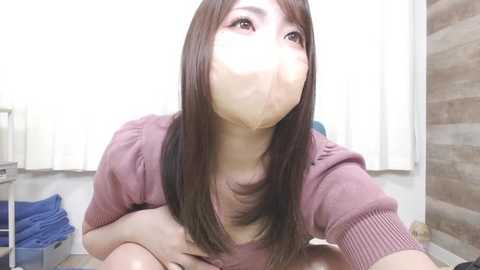 Video of a young Asian woman with straight, shoulder-length brown hair, wearing a pink sweater, with a white face mask covering her mouth and nose. She is indoors, sitting on a tiled floor with a white wall and brown brick wall in the background.