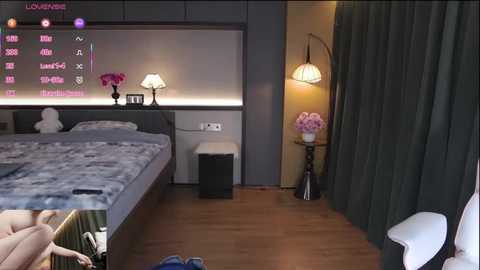 Video of a modern bedroom with a large bed, dark curtains, a wall-mounted TV, a small side table with a vase of pink flowers, and a cozy lamp.