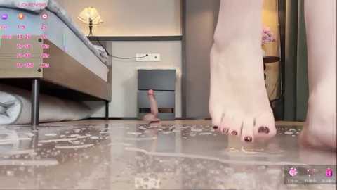 Video of a foot with dark-painted toenails standing in a flooded room, water stains on the floor, bed with blue sheets in the background, and a lamp on the nightstand.