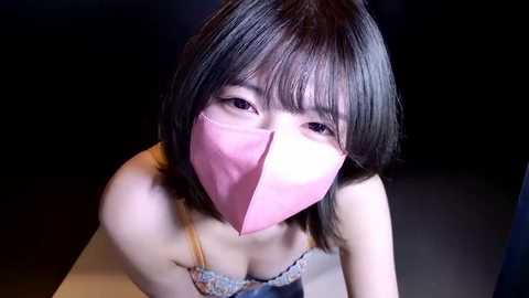 Video of a young Asian woman with short black hair, wearing a pink mask over her face, and a lace-trimmed bra, against a dark background.