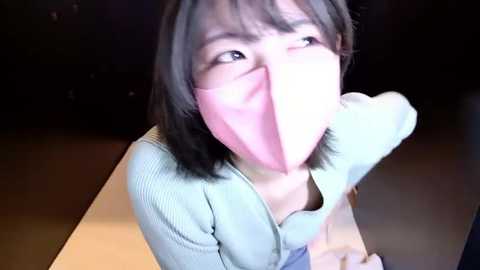 Video of an East Asian woman with straight black hair, wearing a light blue sweater, and a pink mask covering her mouth, looking upward in a dimly lit room.
