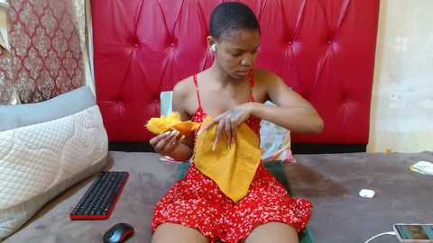 Video of a young African-American woman with short hair in a red floral dress, sitting on a carpeted floor, sewing a yellow fabric piece.