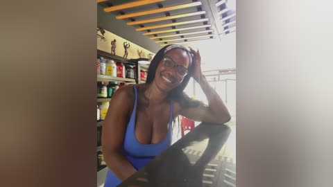 Video of a smiling, dark-skinned woman with long braids in a blue tank top, leaning on a counter in a modern, brightly lit room with wooden beams and shelves of colorful products in the background.