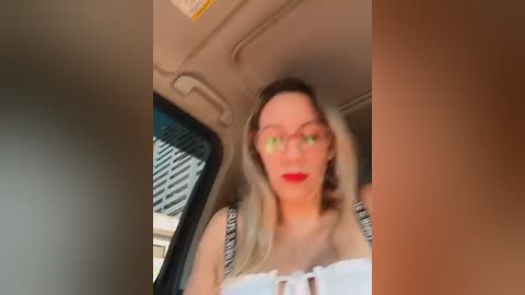 Video of a blonde woman in glasses, wearing a white top, sitting in a car with a blurred cityscape background.