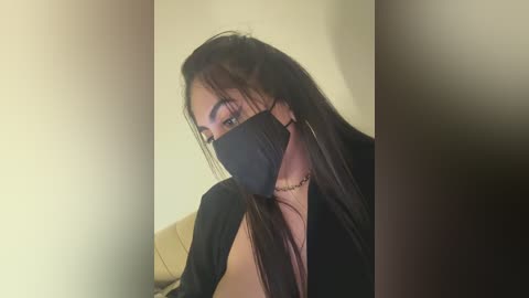 Media: A video of a young woman with long, straight black hair, wearing a black face mask and a black top, seated against a beige wall.