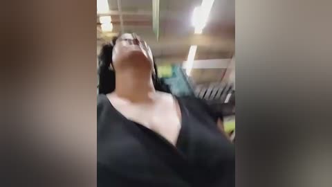 A video of a woman with medium skin tone and curly black hair, wearing a black V-neck sweater, standing with eyes closed, mouth open, in a dimly lit room with fluorescent lights and blurred objects in the background.