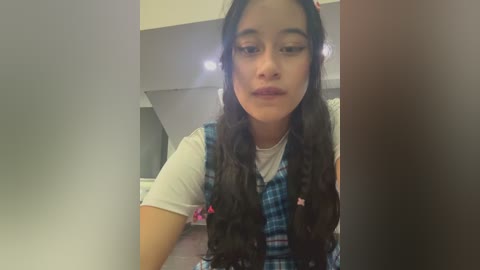 Media: Video of a young Latina woman with long, dark hair, wearing a white T-shirt under a blue plaid vest, indoors, looking down, slightly blurry background.