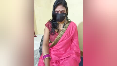 Video of a woman in a bright pink sari with gold embroidery, wearing a black face mask, seated indoors with a beige wall and white bed in the background.