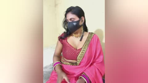 A video of a South Asian woman in a pink sari with gold embroidery, wearing a black face mask, seated indoors with a blurred background.