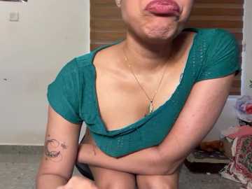 Video of a light-skinned, petite woman with small breasts, wearing a green crop top, kissing her cheek, revealing a tattoo on her arm, in a dimly lit bedroom with a wooden blind.