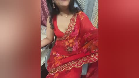 A video of a woman in a vibrant red saree adorned with intricate floral embroidery, seated in a room with patterned walls and a partially visible bed.