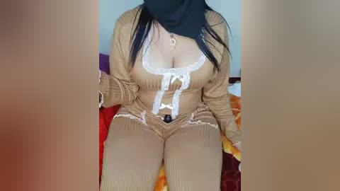 A video of a curvaceous woman with long black hair, wearing a revealing beige jumpsuit with a white lace top, sitting on a bed with a colorful blanket.