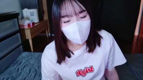 A video of an Asian woman with straight black hair, wearing a white T-shirt with red \"Night\" text, and a white surgical mask, sitting indoors with a wooden dresser and bed in the background.