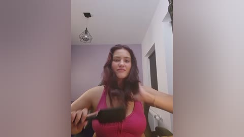 Video of a woman with long, wavy dark hair, wearing a pink tank top, holding a hairbrush, standing in a narrow hallway with white walls and a modern pendant light.