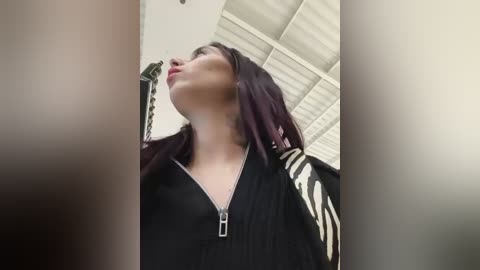 Media: A video of a young woman with fair skin, dark hair, and a black jacket with a zipped neckline. She stands indoors with a modern, white ceiling. The image is slightly blurred, emphasizing a sense of depth.