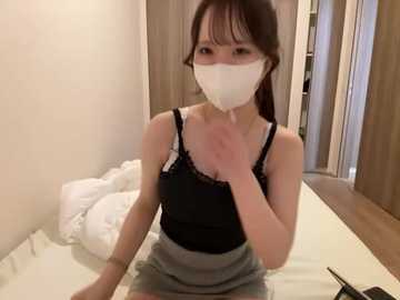 Video of an Asian woman with long black hair and light skin, wearing a black lace-trimmed tank top, gray skirt, white mask, and white gloves, sitting on a white bed in a small, sparsely furnished room with wooden floors.
