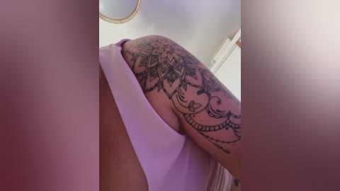 Video of a person with a large, intricate black tattoo on their upper arm, featuring floral and geometric patterns. The background is a blurred indoor setting with a light-colored wall and a circular mirror.