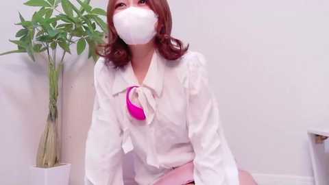 Video of an Asian woman with fair skin and shoulder-length auburn hair, wearing a white blouse with a pink bow, face mask, and sitting on a white floor, surrounded by a potted plant.
