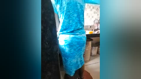 Video of a person in a bright blue tie-dye dress, standing in a kitchen with white tiled walls, a sink, and a blender on the counter.