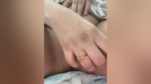 Video of a close-up of a person's hand with a gold ring on their finger, resting on their genital area, which is partially visible. The background shows light-colored fabric.