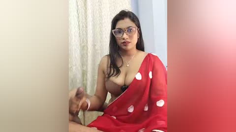 Media: A video of a South Asian woman with medium skin tone and dark hair, wearing glasses, a red sari with white polka dots, and a black bra, standing against a beige curtain, holding a penis.