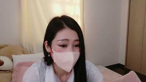 Video of an East Asian woman with long black hair, wearing a white face mask, a white shirt, and red earrings, sitting on a bed with beige and white bedding.