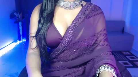 Video of a woman in a deep purple saree with a sheer, embroidered border, adorned with a silver necklace and matching bangles. Background features a modern, dimly lit room with a blue hue.
