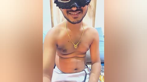 Media: Video of a smiling, shirtless, light-skinned man with a medium build, wearing a black mask, gold necklace, and white shorts, indoors with a bed and pillow in the background.
